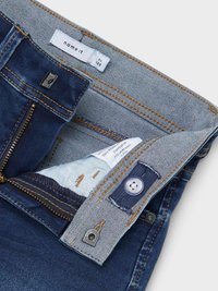 Denim jeans with a dark blue color, gold stitching, metal zipper, and button closure. The waistband features a brand tag and inner fabric detail.
