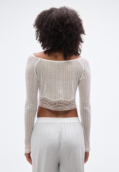 Λευκό crop top από δαντέλα με μακριά μανίκια, με λεπτό λουλουδάτο σχέδιο και γαμπαρωτή τελείωμα. Συνδυασμένο με παντελόνια με ελαστική ζώνη σε ανοιχτό γκρι χρώμα.