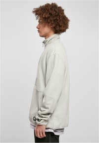 Lys grå fleece-pullover med høy krage, lange ermer og detaljer langs sidesømmen. Har et logo-merke på nedre venstre side.