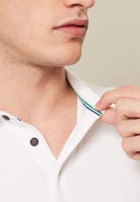Camisa polo blanca con botones negros, que presenta un cuello con rayas multicolores en verde, azul y púrpura. Textura suave, diseño casual.