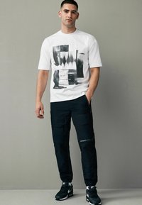 Hvid grafisk t-shirt med sort abstrakt mønster, parret med sorte cargo bukser med lommer og elastiske manchetter, båret med sorte sneakers.