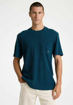 Jonge man met kort haar, gekleed in een donkerteal ribgebreid T-shirt met korte mouwen en borstzak, en beige broek, staand met één hand in zijn zak.