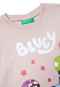 Světle růžové dětské tričko s nápisem "Bluey", malými červenými hvězdami a částečně zobrazenou barevnou kreslenou postavičkou pod nápisem.