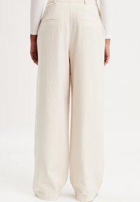 Pantalon à jambes larges en tissu beige clair, avec deux poches arrière et une texture lisse. La ceinture est épurée et au design minimaliste.