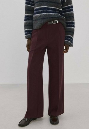 Persona con suéter peludo de rayas oscuras, pantalones anchos color marrón oscuro con cinturón negro, y mocasines marrón oscuro sobre un suelo liso.