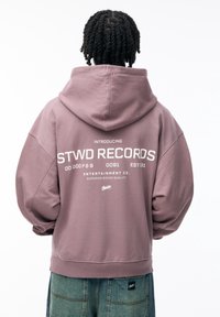 Lila übergroße Kapuzenpullover mit weißem Text auf der Rückseite, der "STWD RECORDS" sagt, aus weichem Stoff gefertigt und mit Kapuze.