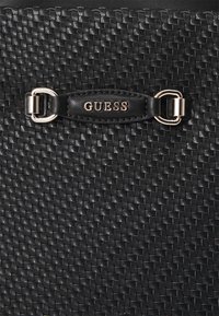 Zwarte geweven handtas gemaakt van textuurmateriaal, met een goudkleurig hardwareaccent en een zwarte leren label met het geperste "GUESS" logo.