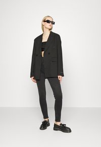Blazer oversized preto com botões duplos, combinado com um top cropped preto, jeans escuros e sapatos pretos com atacadores. Os acessórios incluem óculos de sol quadrados.