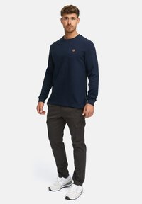 Marine crewneck sweatshirt med tekstureret stof, ribbede ærmer og et cirkulært logo. Sammen med mørke cargo bukser og hvide sportssko.