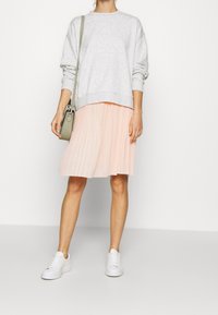 Felpa grigia con maniche lunghe, gonna pesco plissettata e sneakers bianche. Porta una piccola borsa verde. Outfit casual su uno sfondo neutro.