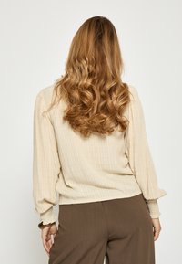 Žena s dlhými vlnitými červeno-hnedými vlasmi oblečená v béžovej blúzke s dlhými rukávmi a hnedých nohaviciach, stojaca chrbtom ku kamere.