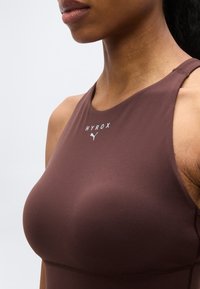 Κοντινό πλάνο γυναίκας που φοράει καφέ αθλητικό τοπ χωρίς μανίκια με εφαρμοστή γραμμή, με τα λογότυπα "HYROX" και Puma στο στήθος.