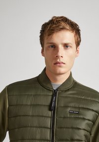 Veste bomber verte olive avec texture matelassée, fermeture éclair et patch logo. Équipée d'un col côtelé ; le tissu semble lisse et léger.