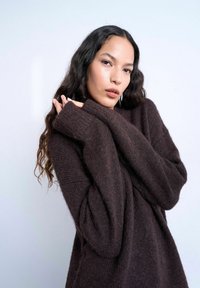 Femme aux longs cheveux bouclés et foncés portant un pull oversize marron foncé, posant avec les mains près du visage sur un fond clair uni.