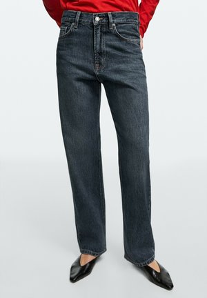 Personne portant un jean gris foncé taille haute et jambes droites, associé à des chaussures noires à bouts pointus et un haut rouge à manches longues.