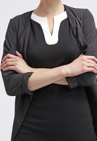 Czarna sukienka z białym detalem przy kołnierzyku, nałożona na szary otwarty żakiet. Materiał wydaje się gładki, z podwiniętymi rękawami. Ręce skrzyżowane.