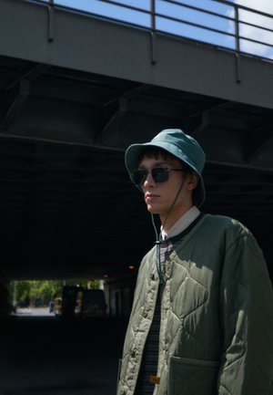 Green quilted jacket with a textured finish, worn over a striped shirt. A teal bucket hat and dark sunglasses complete the look.