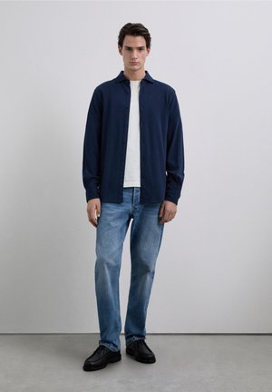 Donkerblauw shirt met lange mouwen, wit shirt eronder, gecombineerd met lichtblauwe jeans en zwarte schoenen. Soepele stof, standaard kraag, knoopsluiting.