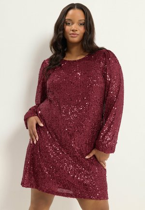Femme portant une robe bordeaux à manches longues avec des sequins et un col rond, posant les mains sur les hanches devant un fond uni.