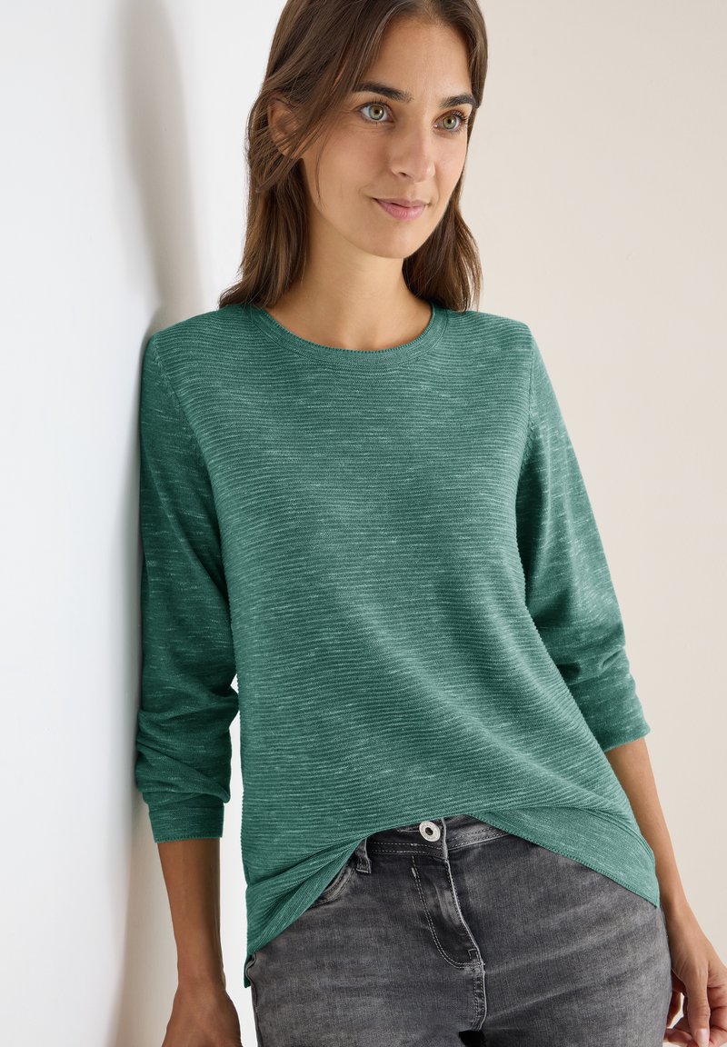 Türkisfarbiges Langarmshirt mit Rundhalsausschnitt und strukturierter, gestreifter Musterung, kombiniert mit grauen Jeans mit Knopfverschluss.