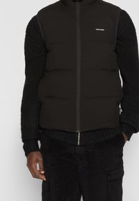 Schwarze gepolsterte Weste mit hohem Kragen und Frontreißverschluss, getragen über einem strukturierten schwarzen Strickpullover und Cordhosen. Dezente Markenkennzeichnung sichtbar.