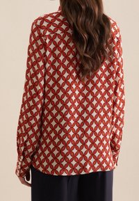 Kvinna med långt vågigt brunt hår bär en vit blus med ett mönster av en orange länkad kedja, i kombination med mörka byxor, visas från ryggen.