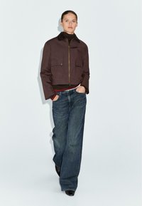 Braune, kurze Jacke mit einem braunen Kragen, zwei Vorderstaschen, kombiniert mit weiten blauen Jeans. Glatte Textur mit einem Reißverschluss auf der Vorderseite.