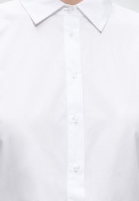 Gros plan sur une chemise blanche boutonnée avec un col classique et cinq boutons visibles au centre devant.