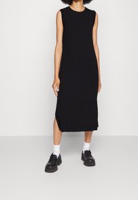 Robe noire sans manches à côtes avec encolure ronde, longueur genou, avec des fentes latérales. Portée avec des chaussettes blanches et des chaussures noires épaisses.