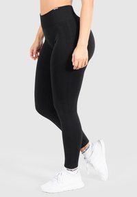 Leggings neri a vita alta realizzati in tessuto elasticizzato, con una texture liscia e design senza cuciture, abbinati a scarpe sportive bianche.