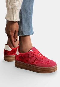 Baskets en velours rouge avec une tige texturée, accents blancs et épaisse semelle en caoutchouc. Présente des lacets roses et un design rayé sur le côté.
