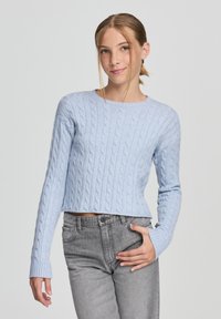 Maglione azzurro chiaro a trecce con scollatura tonda e maniche lunghe, abbinato a jeans grigi a vita alta, caratterizzati da una sottile trama e dettagli.