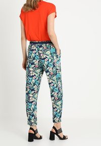 Kobieta ubrana w czerwony krótki rękaw z niebieskimi spodniami w kwiaty i czarne sandały na wysokim obcasie z otwartymi palcami, stoi z rękami w kieszeniach.