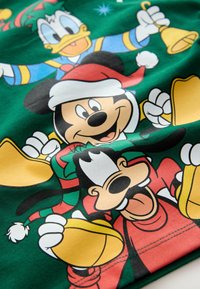 Žalias audinys su animacinių filmų herojais: Mikis Pūkuotukas, Donaldas Pūkuotukas ir Gufis, apsirengę spalvingais šventiniais drabužiais, su varpeliais ir šventinėmis detalėmis.