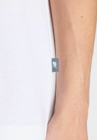 Valkoinen puuvillainen t-paita lyhyillä hihoilla, jossa on pieni harmaa logo-etiketti sivusaumassa ja suora helma. Siisti tekstuuri, rento muotoilu.