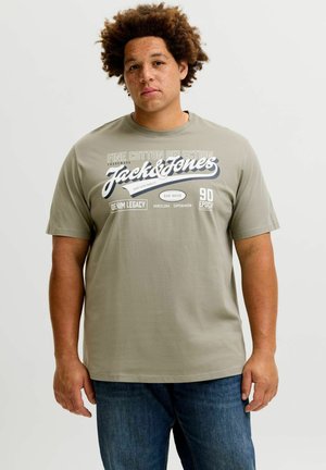 Mann mit lockigem Haar und Sommersprossen, der ein grünes Jack & Jones T-Shirt und blaue Jeans trägt, steht vor einem einfarbigen Hintergrund.