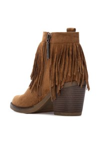 Stivale alla caviglia in suede marrone con dettagli in frange, zip laterale e tacco in legno impilato. Superficie testurizzata con una punta arrotondata.