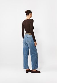 Kobieta z włosami upiętymi w bun, ubrana w brązową bluzkę z długim rękawem, niebieskie spodnie typu palazzo z paskiem w panterkę i brązowe mokasyny, stoi na tle białej ściany.