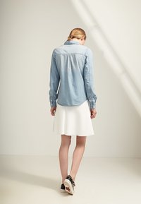Lyseblå denimskjorte med lange ærmer, buet kant og knaplukning ved håndleddene, parret med en hvid nederdel og sorte sneakers.