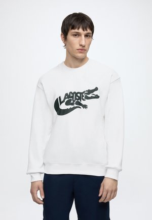 Jeune homme portant un sweat-shirt blanc avec un grand logo crocodile Lacoste noir et un pantalon bleu marine, debout sur un fond blanc uni.