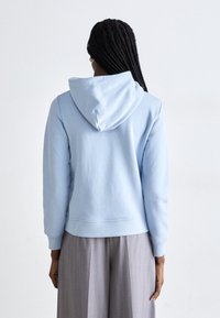 Ανοιχτό μπλε φούτερ με κουκούλα και χαλαρή εφαρμογή, διαθέτει τσέπη καγκουρό και ριπές στους μανσέτες. Λείος υφασμάτινος χαρακτήρας, μινιμαλιστικός σχεδιασμός.