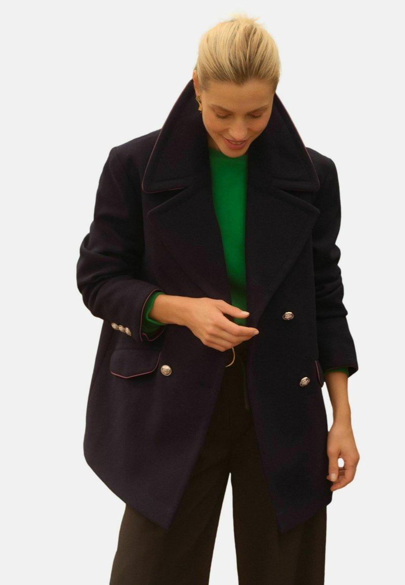 Dvojradový námorní plášť s veľkým golierom, striebornými gombíkmi a kontrastným ružovým lemovaním. Nosený cez zelený top, má voľný strih.