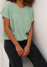 Hellgrüne kurzärmlige Bluse mit glatter Textur, runder Ausschnitt und dezenten vertikalen Streifen, kombiniert mit dunkelgrauen Jeans.