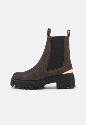 Bota Chelsea de ante marrón oscuro con paneles elásticos negros en los lados, tiradores, suela gruesa con tacos y un tacón bajo y robusto sobre un fondo blanco.