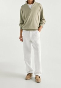 Licht olijfkleurig pullover met een halvemaanrits kraag, gecombineerd met witte broek en beige veterschoenen. Materialen lijken zacht en gestructureerd.