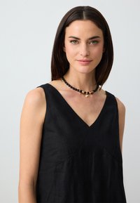 Frau mit glatten, dunklen Haaren, die ein ärmelloses schwarzes Kleid und eine schwarze Perlenkette mit goldenen Muschelanhängern vor einem hellen Hintergrund trägt.