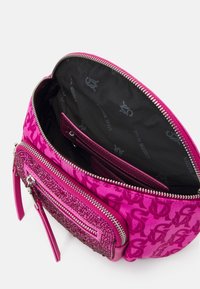 Vaaleanpunainen käsilaukku, jossa on teksturoitu pinta, vetoketjukiinnitys, kimaltelevia yksityiskohtia ja logokuosiset sisävuoret. Kompakti muotoilu.