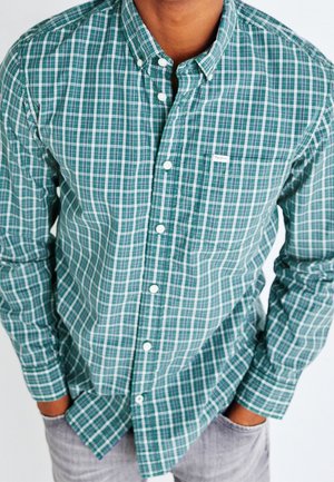 Hombre con camisa abotonada de cuadros verdes y blancos, con las manos en los bolsillos de pantalones gris claro, mostrado desde el cuello hasta las caderas.
