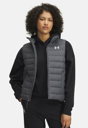 Pohodlná šedá vesta s výplňou a zipsom vpredu, vysokým golierom, nosená cez čierny mikinový sveter. Obsahuje horizontálne prešívanie a logo Under Armour na hrudi.