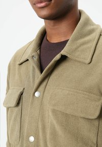 Olijfgroene fleecevest met een gestructureerde afwerking, voorzien van een omgevouwde kraag, drukknopen en twee borstzakken. Bruine laag eronder.